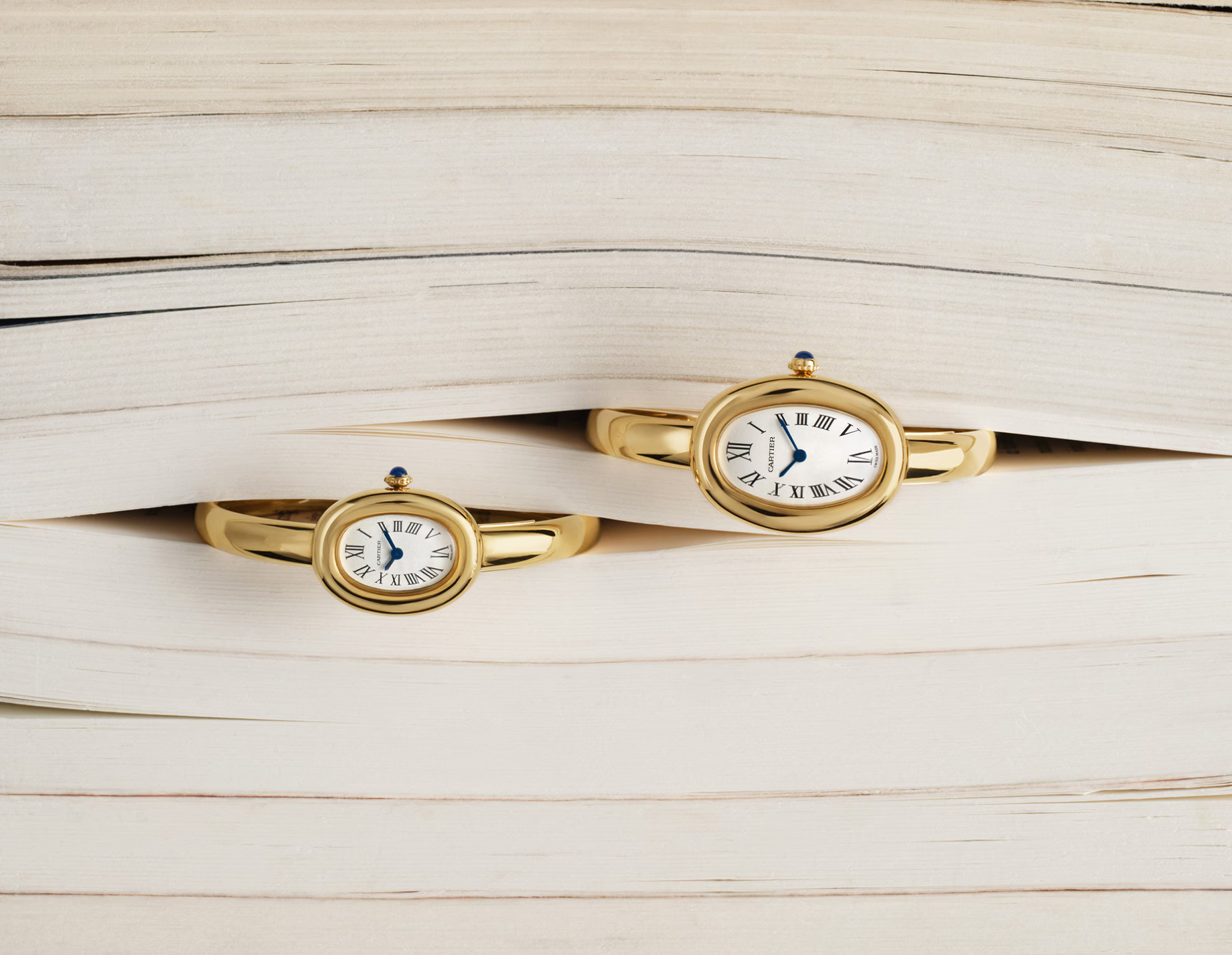 Two gold Cartier wristwatches with white faces and Roman numerals, displayed horizontally between the pages of stacked open books.